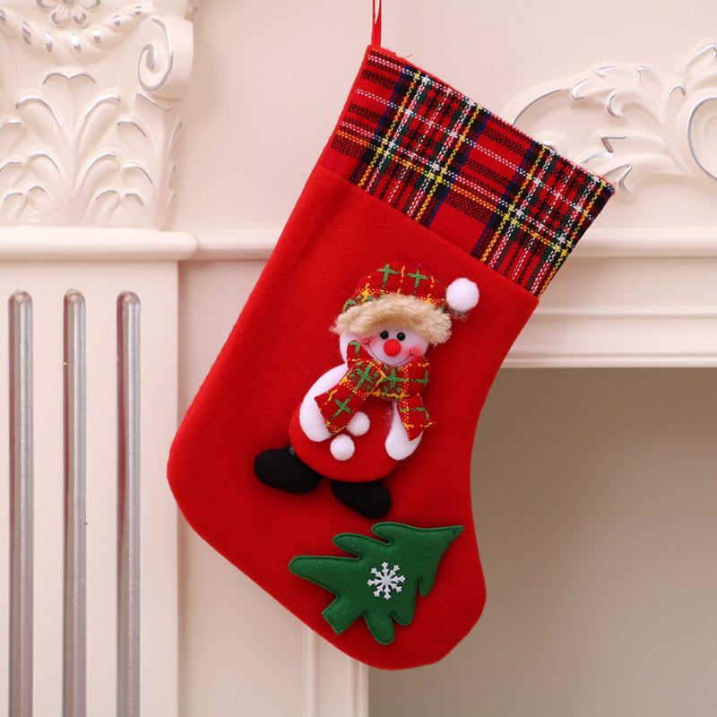 Red Christmas stocking with plaid cuff, Santa Claus decoration, and green Christmas tree applique hanging on a white mantle.